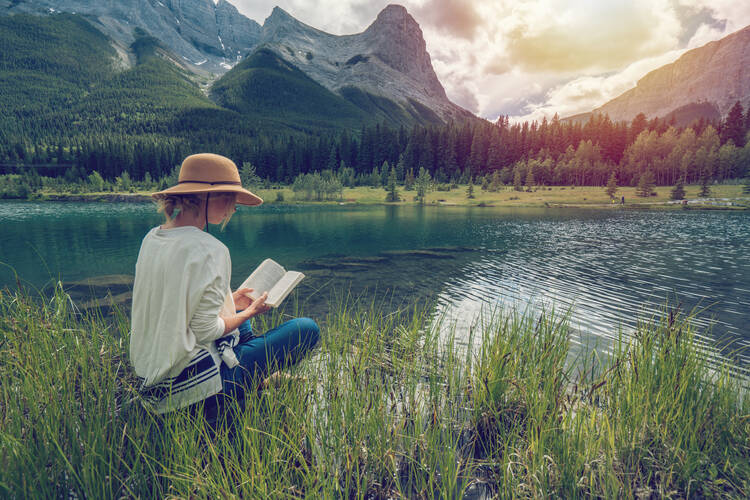 A woman reads a book while sitting on the bank of a calm river, with mountains in the background. (iStock/swissmediavision)